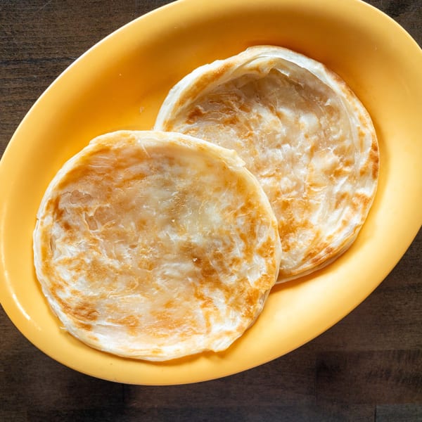 Two golden-brown paratha flatbreads are served on a bright yellow oval plate on a wooden table.