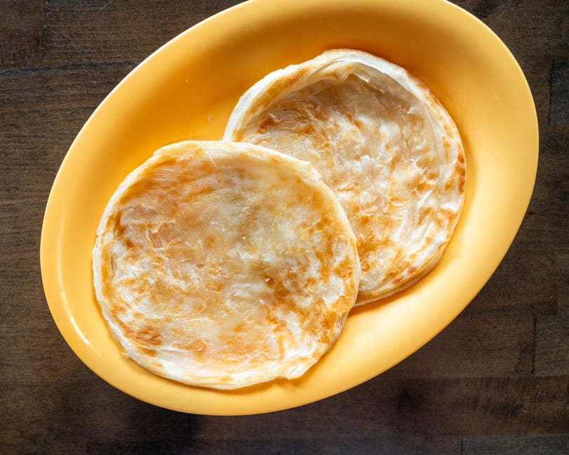 Two golden-brown paratha flatbreads are served on a bright yellow oval plate on a wooden table.
