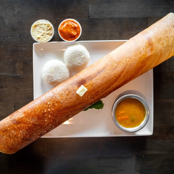 A long, golden-brown dosa with butter, two idlis, sambar, and two chutneys on a white plate.