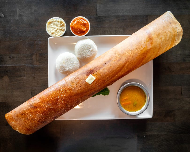 A long, golden-brown dosa with butter, two idlis, sambar, and two chutneys on a white plate.