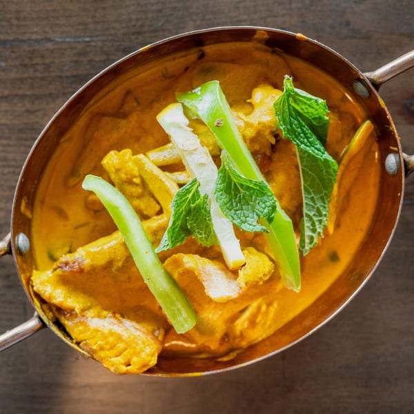 Overhead view of a copper pot filled with yellow curry, garnished with mint, lime, and green peppers.