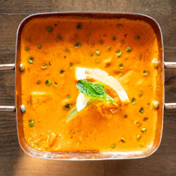Overhead view of a copper pot filled with orange curry, green peas, and lemon slices.
