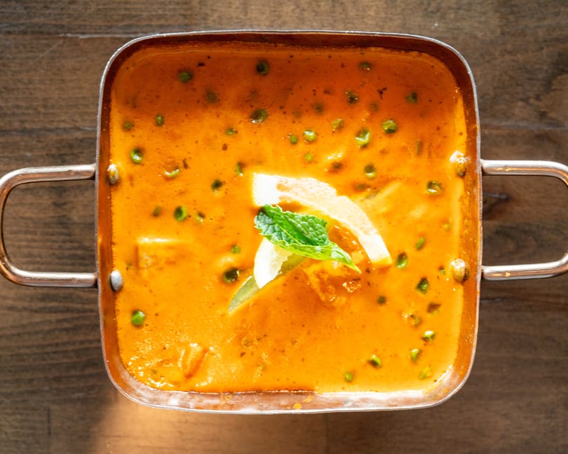 Overhead view of a copper pot filled with orange curry, green peas, and lemon slices.