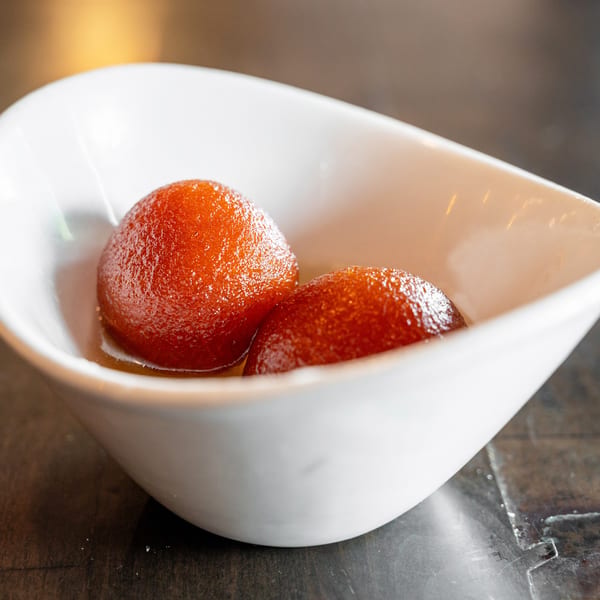 Two glistening gulab jamun in a white, boat-shaped bowl, resting in syrup on a dark surface.
