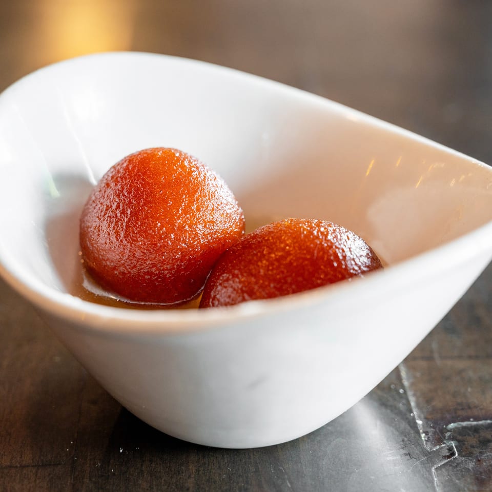 Two glistening gulab jamun in a white, boat-shaped bowl, resting in syrup on a dark surface.