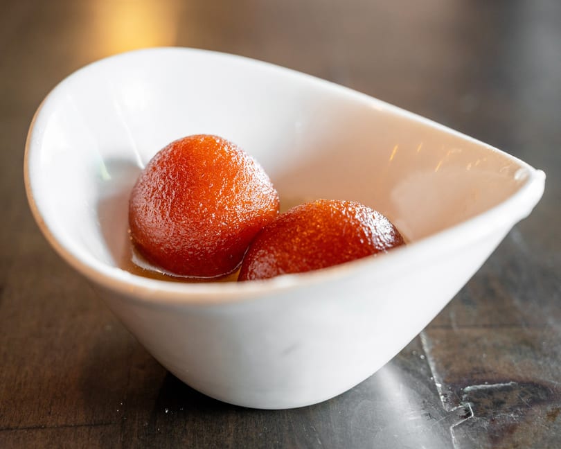 Two glistening gulab jamun in a white, boat-shaped bowl, resting in syrup on a dark surface.