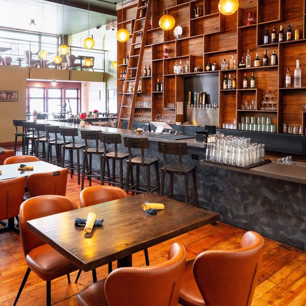 Restaurant interior with a long bar, wooden shelves filled with bottles, and tables with orange chairs.
