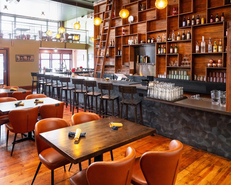 Restaurant interior with a long bar, wooden shelves filled with bottles, and tables with orange chairs.